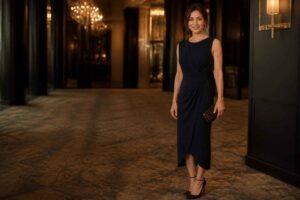 Elegancka kobieta w granatowej sukience stoi w luksusowym hotelowym wnętrzu, trzymając czarną kopertówkę